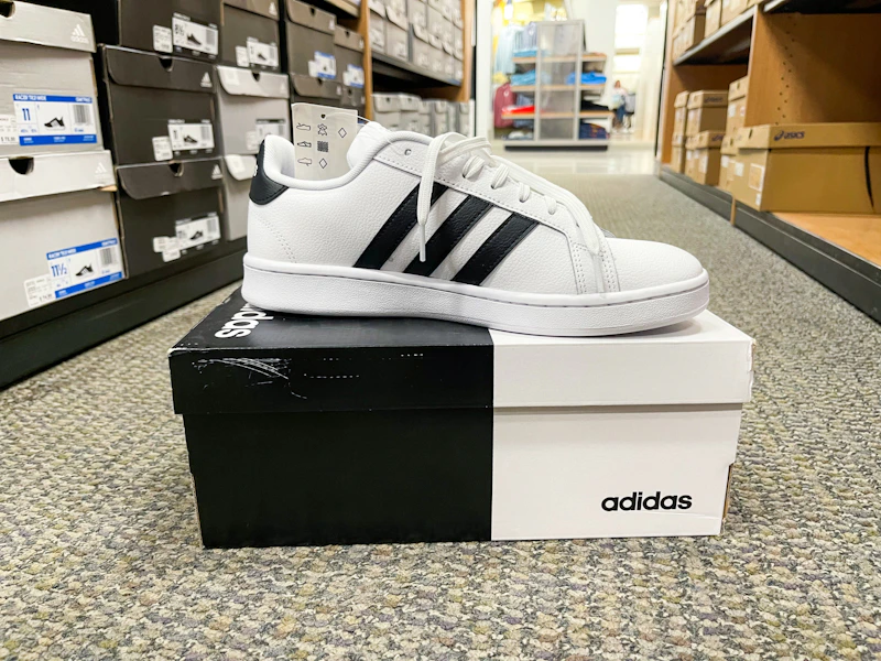 An Adidas Grand Court Mens Sneaker sitting on top of an Adidas box in the middle of a store aisle.
