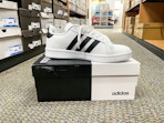 An Adidas Grand Court Mens Sneaker sitting on top of an Adidas box in the middle of a store aisle.