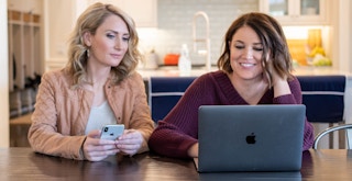 Two woman sitting at a table looking at a laptop computer screen.