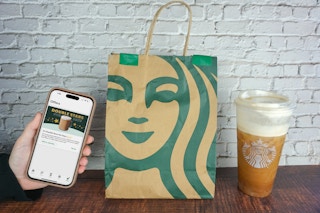 Someone holding a phone displaying the Starbucks Double Stars for Days promotion next to a Starbucks bag and trenta iced coffee