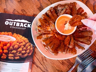 A person reaching their hand in to dip a piece in the Bloomin' Onion sauce cup on a table next to an Outback Steakhouse menu.