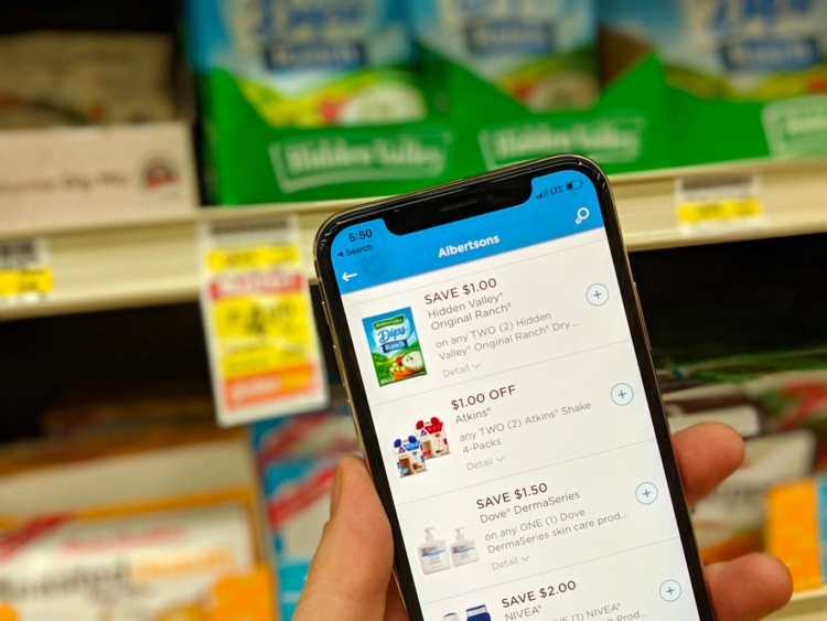 Person holding up a phone showing digital coupons on the Albertsons app, in front of groceries