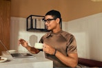 A man wears bluetooth glasses and holds a cup in front of his computer in a kitchen.