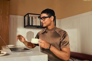 A man wears bluetooth glasses and holds a cup in front of his computer in a kitchen.