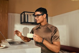 A man wears bluetooth glasses and holds a cup in front of his computer in a kitchen.