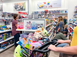 A woman pushing a kohl's cart filled with toys in a busy toy aisle.