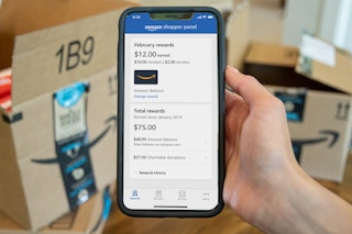 a phone with the amazon shopper panel app on screen being held up in front of amazon boxes