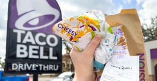 Someone holding up a Crispy Melt Taco and Taco Bell Bag in front of a Taco Bell sign