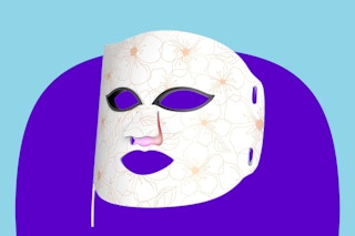 This red light therapy face mask is white with faintly drawn flowers and has eye holes, a nose flap, and a mouth hole plus a cord.