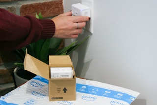 A person plugging a smart plug into an outlet next to a box of more smart plugs on top of an Amazon shipping envelope.