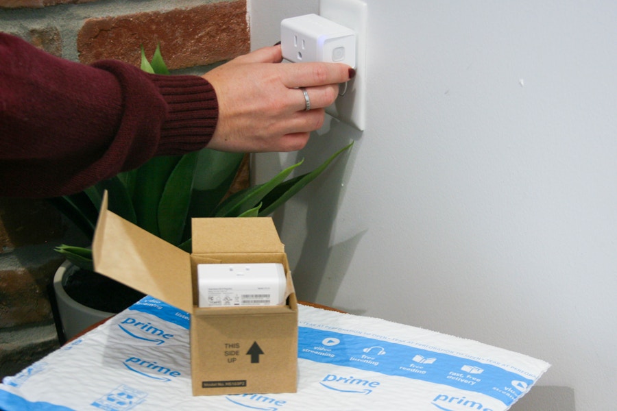 A person plugging a smart plug into an outlet next to a box of more smart plugs on top of an Amazon shipping envelope.