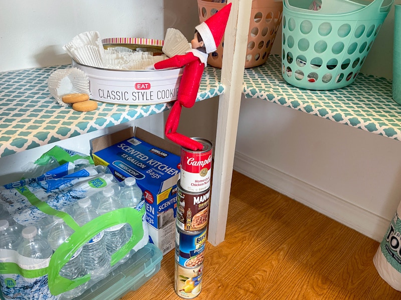 elf on the shelf doll standing on stacked cans getting into a tin of cookie in the pantry 