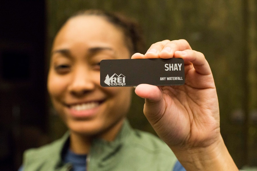 A woman holding up an REI employee name tag that reads "Shay"