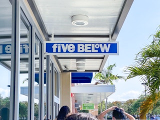 A sign for Five Below hanging from an overhang above the heads of people waiting in a line.