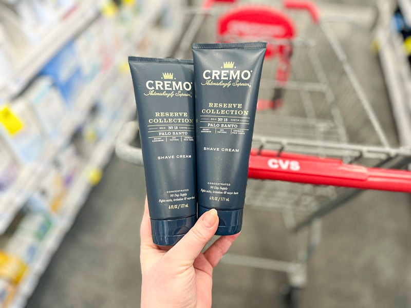 person holding 2 bottles of cremo shave cream over a cvs cart