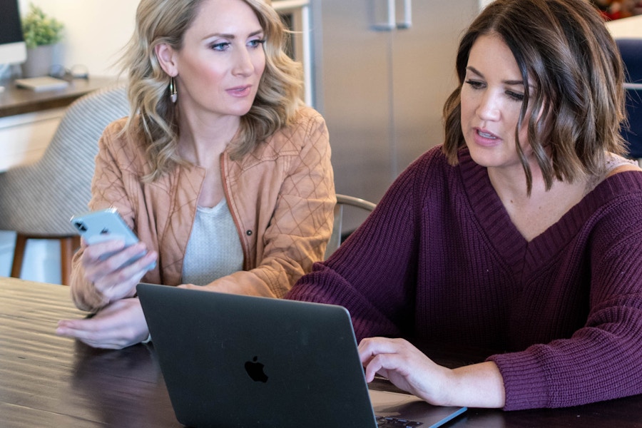 Two woman sitting at a table, talking while one holds a cell phone and the other looks at a laptop.