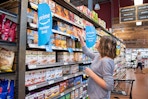 A woman shopping at Whole Foods, reaching up to a shelf tagged with blue Amazon Prime Members Deal signs.