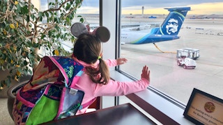 A little girl with Minnie Mouse ears looks out the window at the airport at Alaskan airlines.