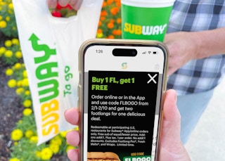 Someone holding a cell phone displaying the Subway app offer for Buy One Get One Free footlong sandwiches from February 1st through the ...