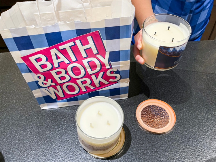 A person holding a 3-wick candle that has been lit before next to a Bath & Body Works bag and an unused 3-wick candle.