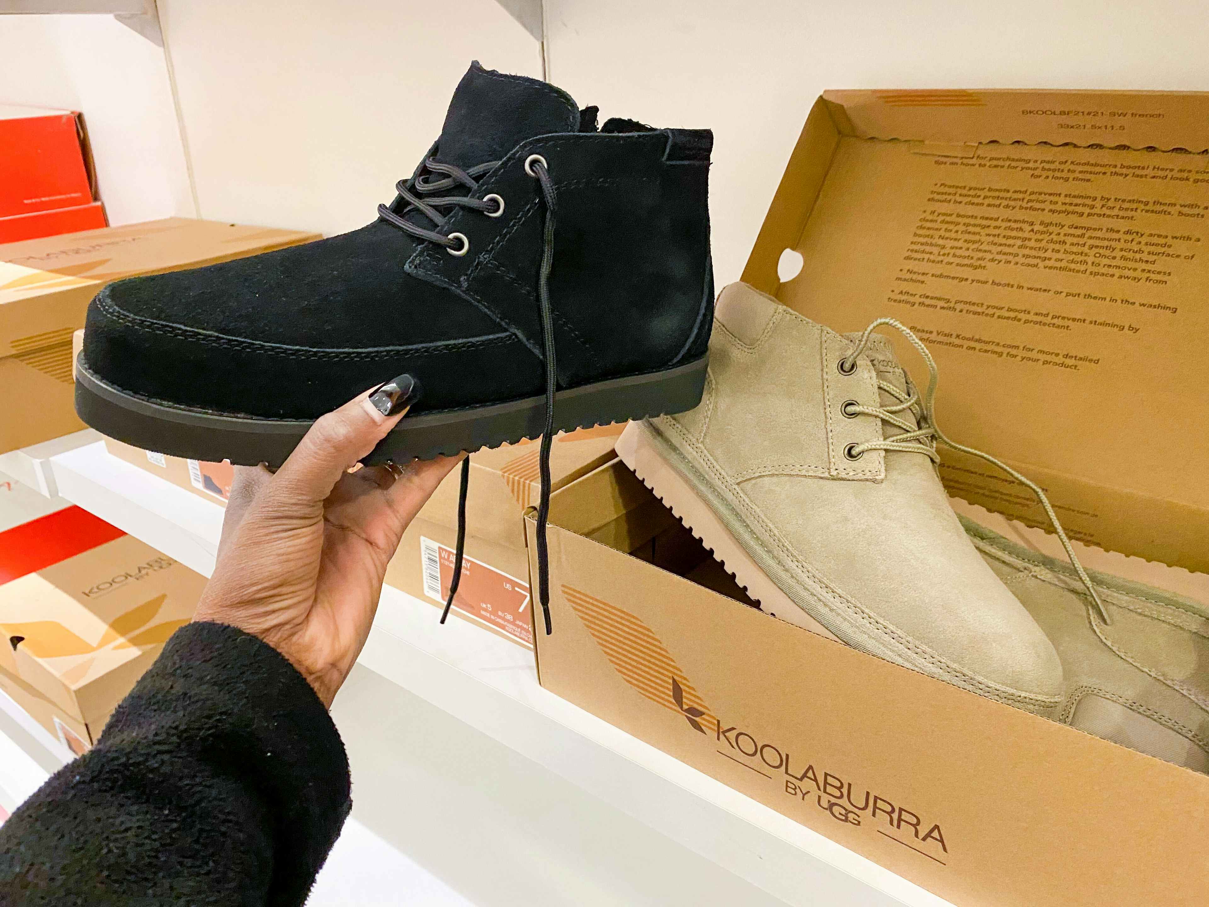 A person holding up a Koolaburra display boot near another pair of Koolaburra boots in their box on a shelf at Macy's.