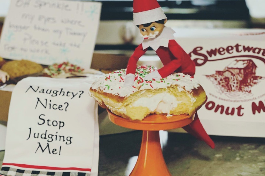 an elf on the shelf pretending to eat a donut next to a funny kitchen towel