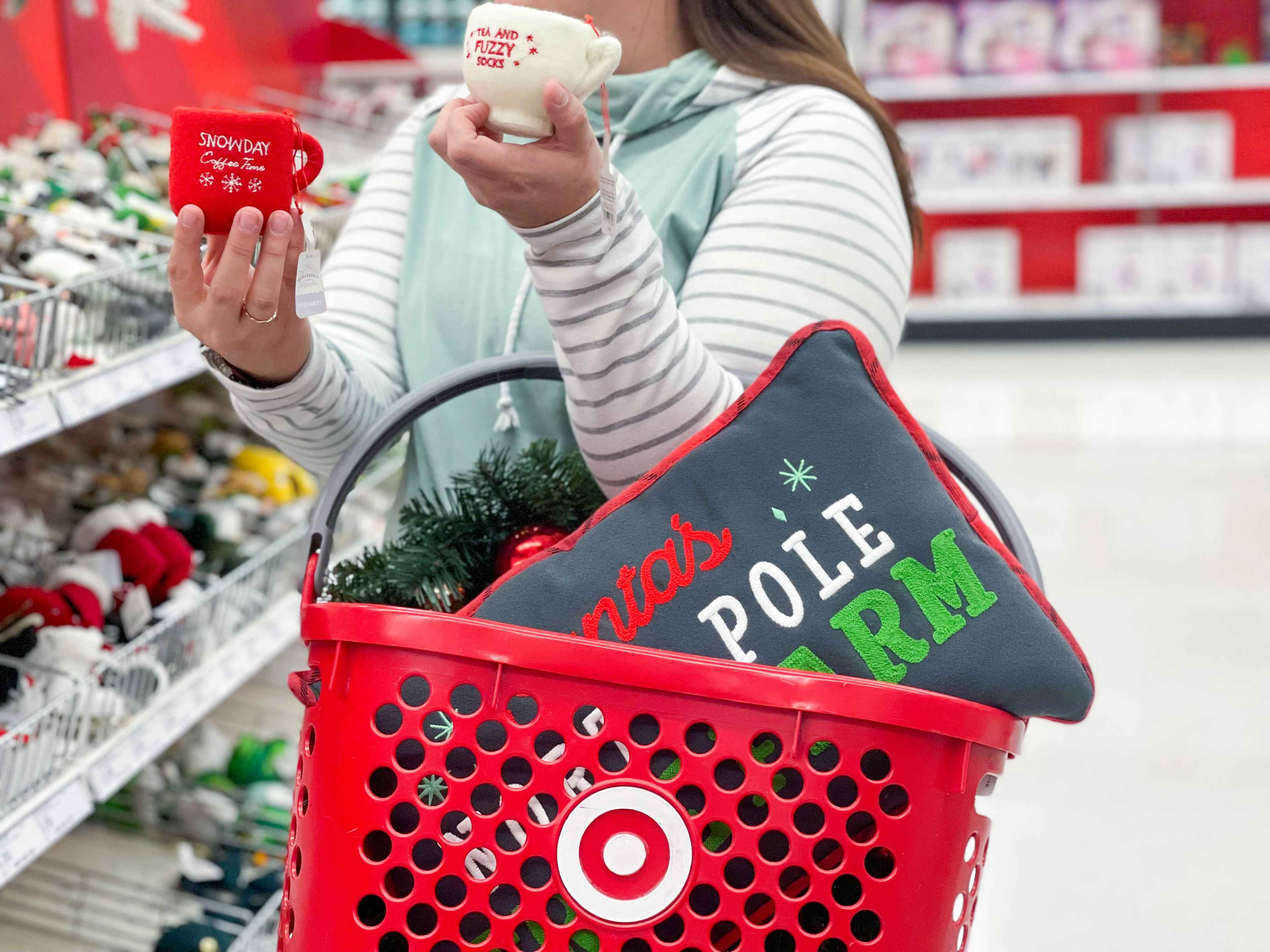 A person carrying a Target hand basket with a holiday-themed pillow in it on her arm, and holding two holiday-themed mugs in an aisle at ...