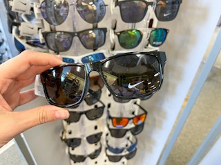 Black Nike sunglasses held up by hand in front of a sunglass stand.