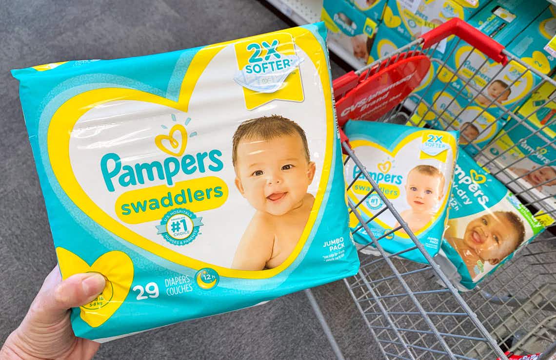 A person's hand holding up a Jumbo Pack of Pamper's swaddlers diapers in front of a shopping cart, with two more packages of diapers, par...