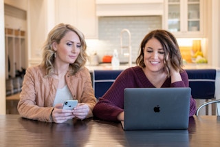 Two woman sitting at a table looking at a laptop computer screen.