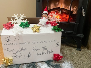 elf on the shelf doll peaking out of a box from the north pole in front of a fake fireplace 