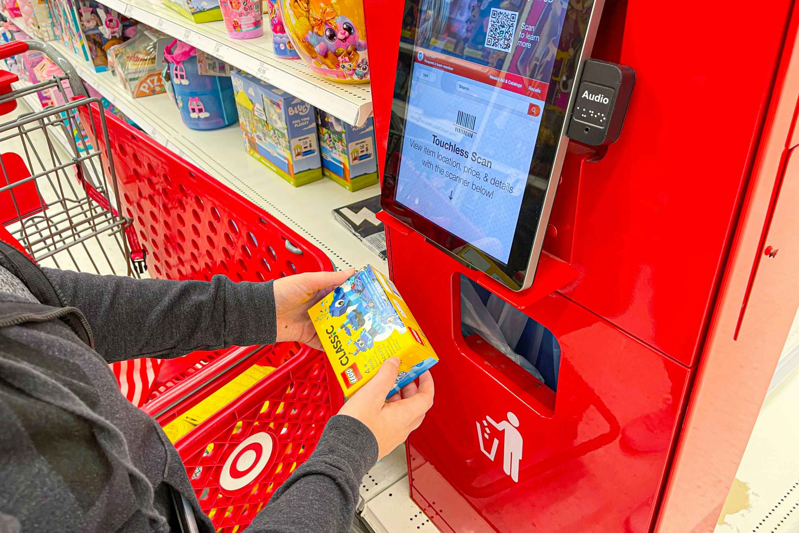 A person standing next to a Target shopping cart, scanning a LEGO classic toy at the price check scanner inside of Target.