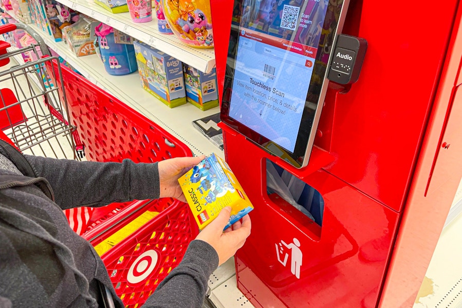 A person standing next to a Target shopping cart, scanning a LEGO classic toy at the price check scanner inside of Target.