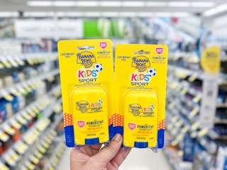 Hand holding two packages of Banana Boat Kids Stick Sunscreen in front of an aisle