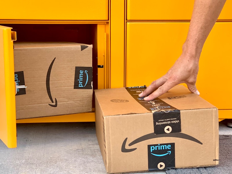 A person's hand going to pick up an Amazon box that is sitting on the ground next to another Amazon box inside an Amazon hub locker.