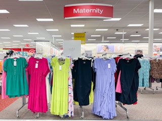 An area shot of a maternity section in a store. Maternity clothing hanging from store racks with a Maternity sign hanging from the ceiling.