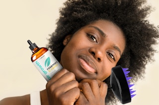 A woman smiles while holding a bottle of Artnaturals Hair Growth Oil and a scalp massager.
