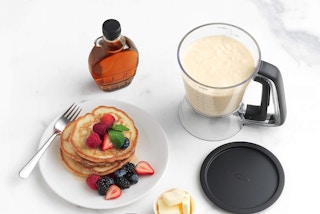 a batter dispenser next to a plate of pancakes and syrup