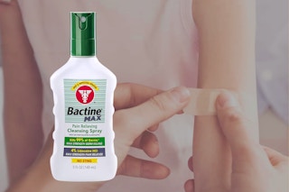 An adult puts a bandage on a child's arm in the background. In the foreground, Bactine Max First Aid Spray.