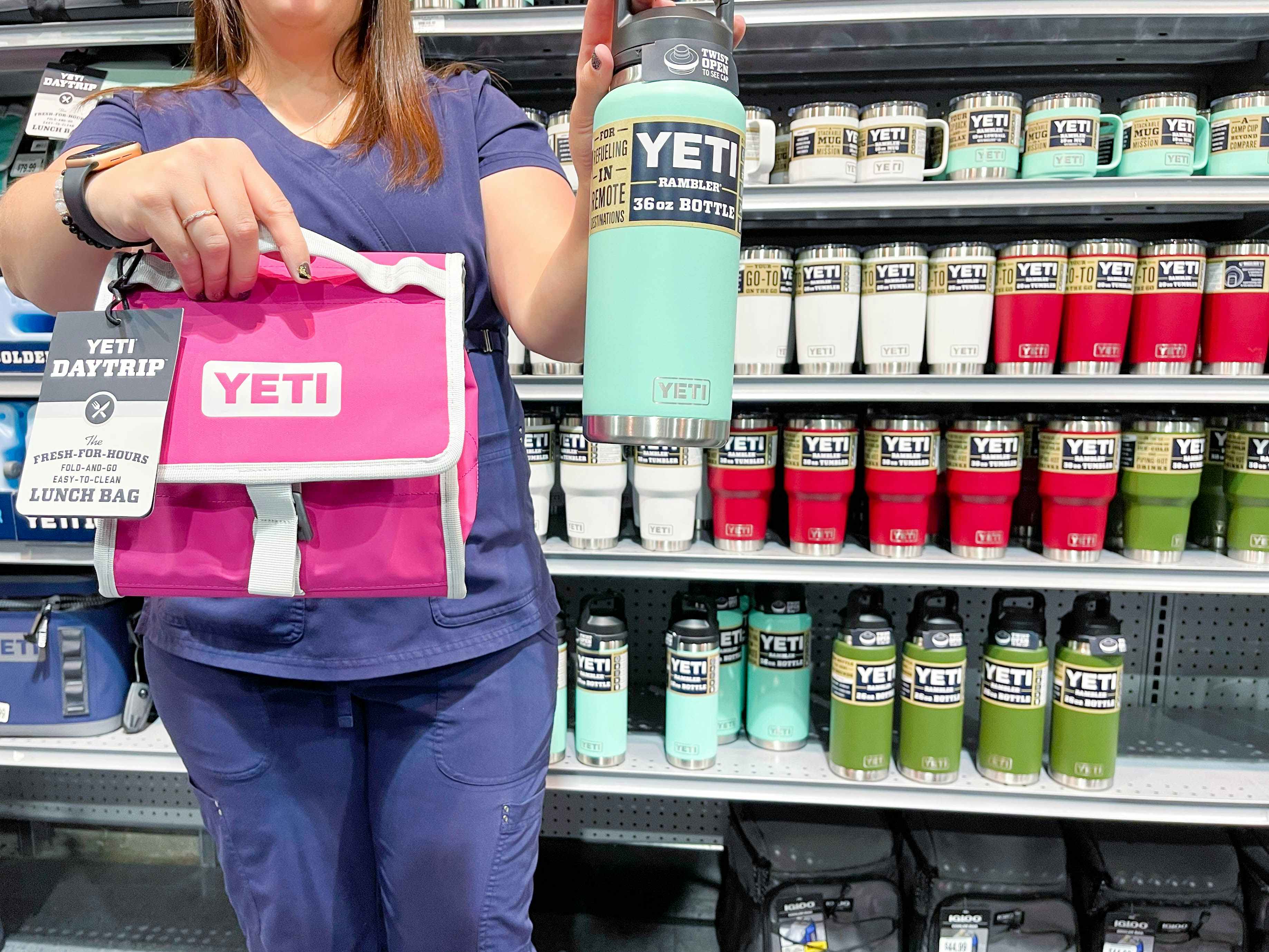 A first responder in scrubs holding a Yeti daytrip lunch bag and a Yeti Rambler water bottle in front of a shelf of more Yeti products.