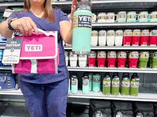 A first responder in scrubs holding a Yeti daytrip lunch bag and a Yeti Rambler water bottle in front of a shelf of more Yeti products.