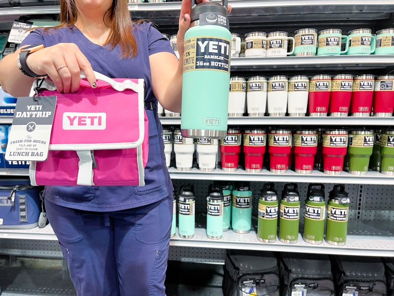 A first responder in scrubs holding a Yeti daytrip lunch bag and a Yeti Rambler water bottle in front of a shelf of more Yeti products.