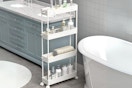 A white wheeled slim storage cart sits in a bathroom with toiletries on it.