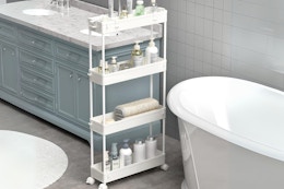 A white wheeled slim storage cart sits in a bathroom with toiletries on it.