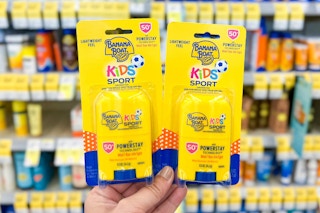 Hand holding to packages of Banana Boat Kids Stick Sunscreen in front of a shelf of sunscreen