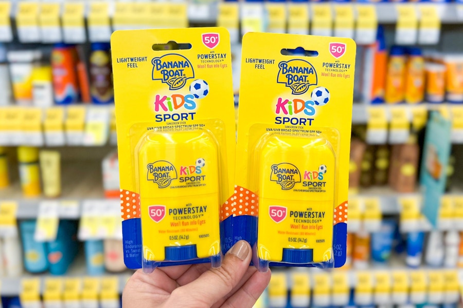 Hand holding to packages of Banana Boat Kids Stick Sunscreen in front of a shelf of sunscreen