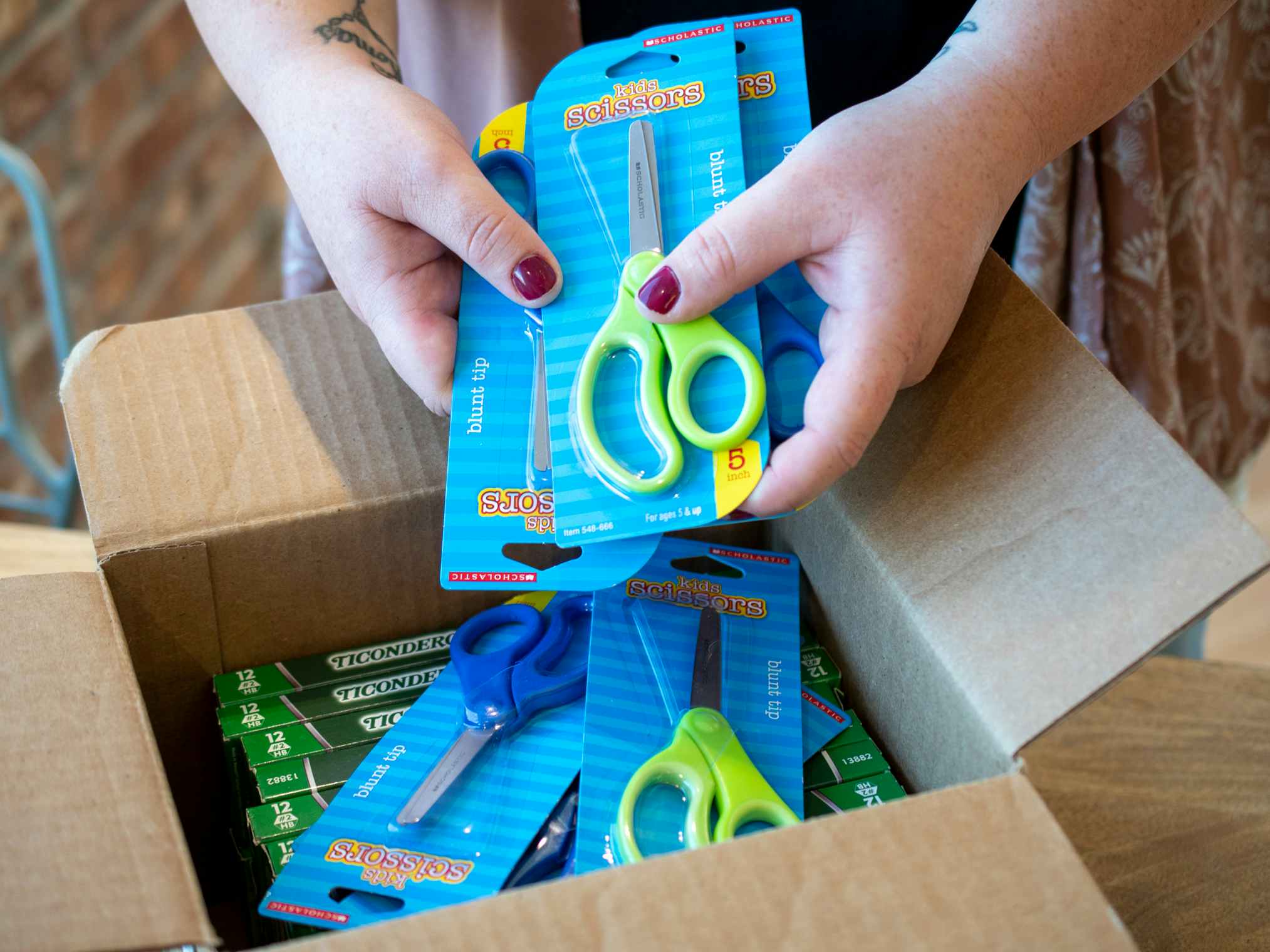 A person taking multiple pairs of scissors out of a shipping box with more scissors and boxes of pencils in it.