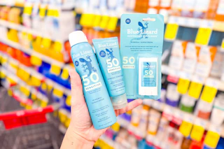 person holding 3 blue lizard sunscreens in front of shelves