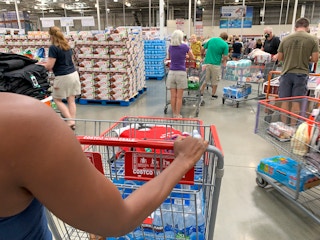 A woman from behind, pushing a Costco shopping cart through a crowd of other shoppers also pushing carts.