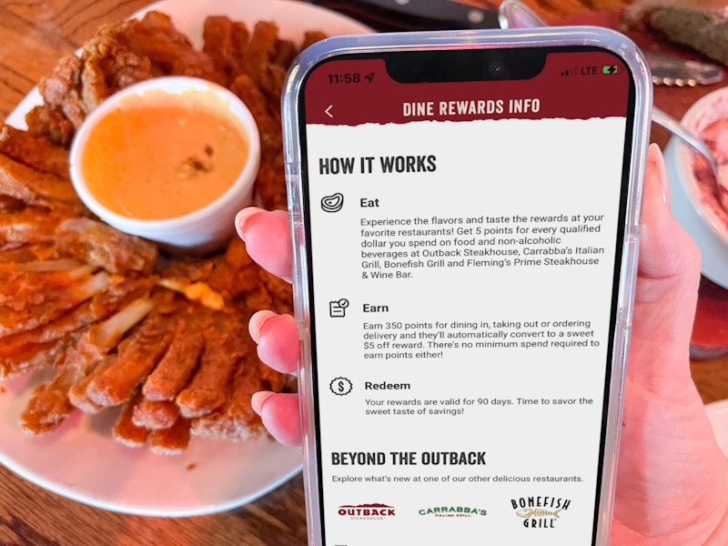 A person's hand holding their phone displaying Outback Steakhouse's Dine Rewards Info in front of a plate of a Bloomin' Onion